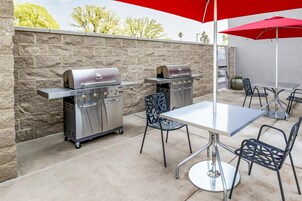 Un patio exterior con dos parrillas de acero inoxidable Char-Broil, una mesa, sillas y un paraguas rojo.