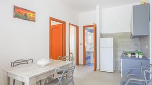 Een compacte keuken met een koelkast, spoelbak en een klein aanrecht met potplanten. Er is een eethoek met een tafel en stoelen, en een deur die naar een andere kamer leidt.