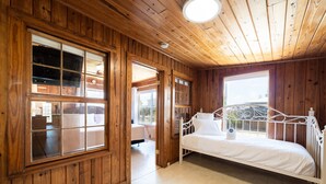 A wooden-paneled room with a white metal bed, large windows, and a ceiling light.
