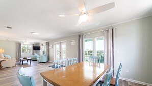 A spacious living room with a wooden dining table, light blue chairs, a ceiling fan, and a television.