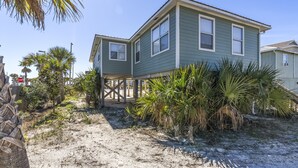 A two-story house with a sandy foundation, surrounded by palm trees and a stop sign in the background.
