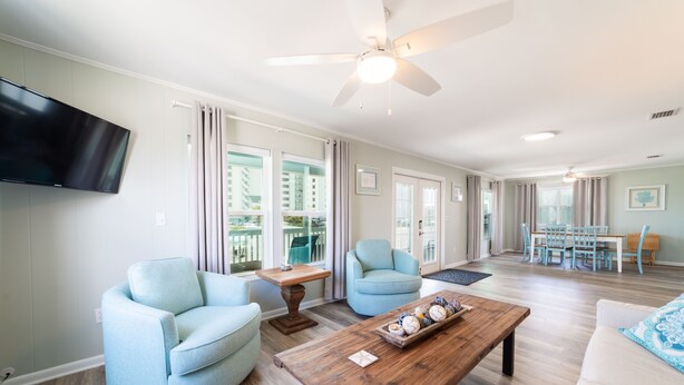 A spacious living room with a ceiling fan, a flat-screen TV mounted on the wall, and a dining area with a table and chairs.