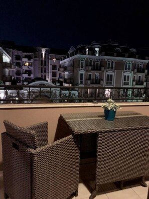 A balcony with wicker furniture and a potted plant, overlooking a row of buildings with balconies. - Luxury Apartment in Balkan Breeze 7 in Sunny Beach (Sunny Beach)