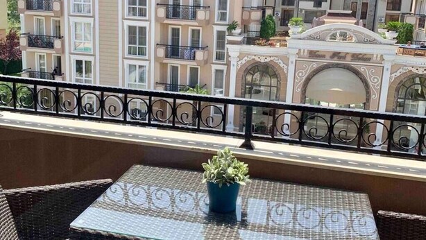 A balcony with wicker furniture and a potted plant, overlooking a row of multi-story buildings with balconies and decorative elements. - Luxury Apartment in Balkan Breeze 7 in Sunny Beach (Sunny Beach)
