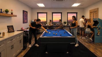 Mesa de billar en una sala con vistas a la puesta de sol a través de amplias ventanas, zona de bar y mostrador con menú.