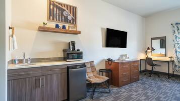 A compact kitchen with a microwave, coffee maker, and a refrigerator. There is a chair, a desk with a computer, and a flat-screen TV mounted on the wall.