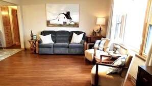 Una sala de estar con un sofá gris, un sillón color beige y piso de madera. Hay un cuadro en la pared y una lámpara sobre una mesita.