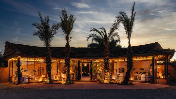 A modern, open-plan villa with a thatched roof, surrounded by palm trees, featuring large glass windows and a well-lit interior. - Casa Abracadabra (Marrakech)