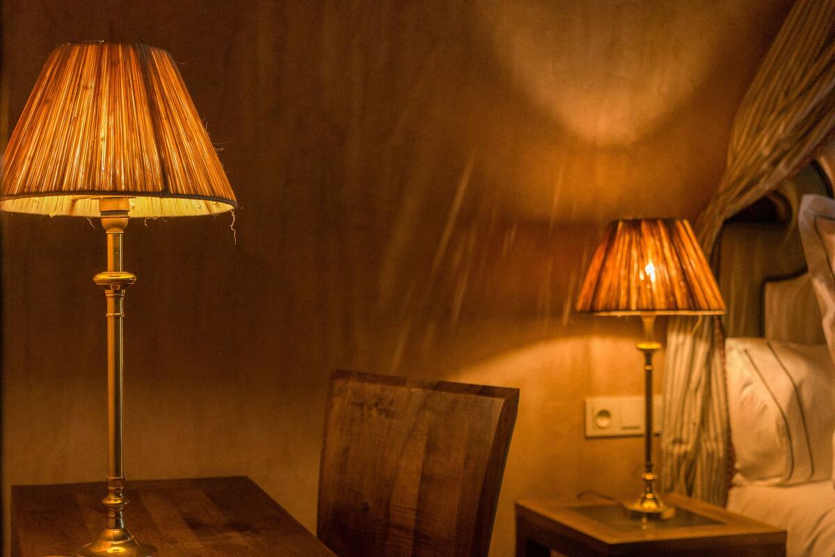 A dimly lit room with a wooden table, two lamps, a chair, and a bed with a headboard.