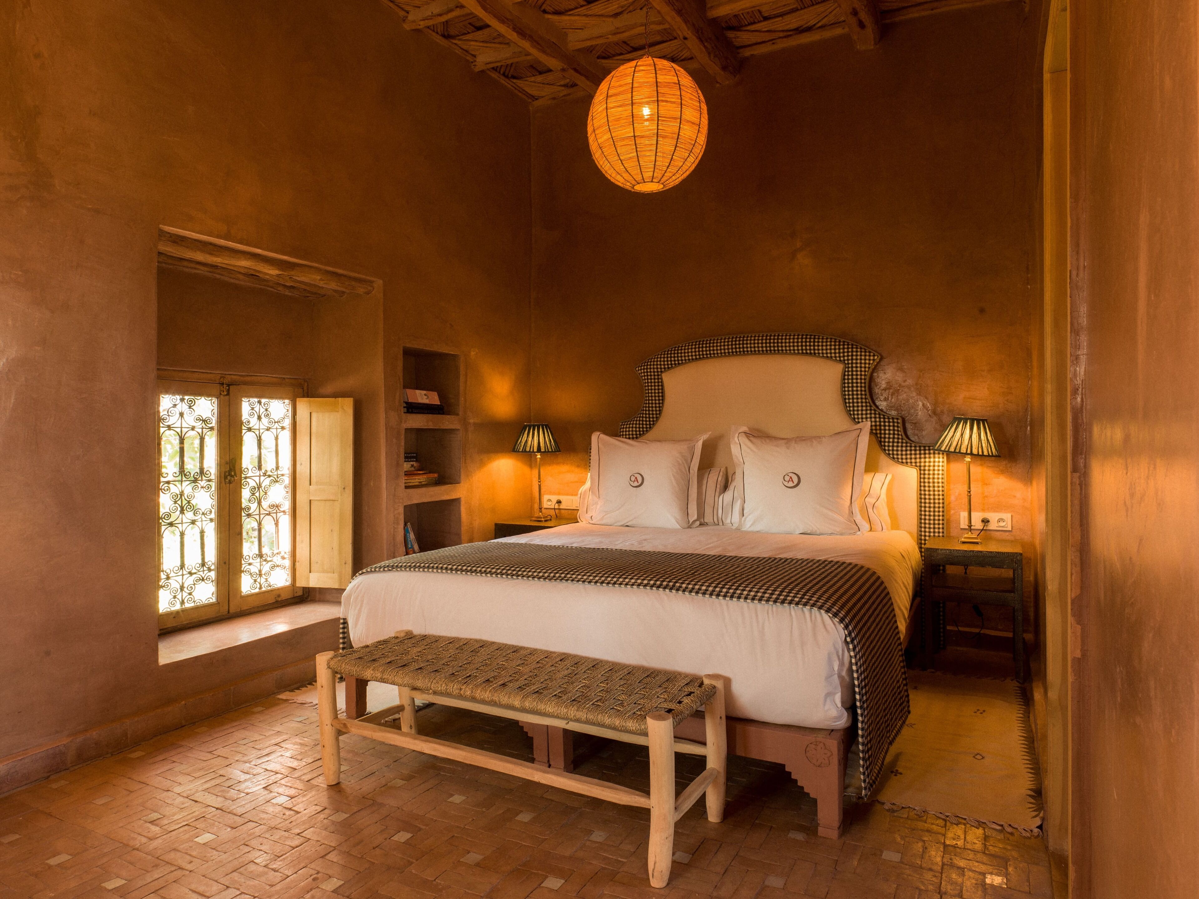 A cozy bedroom with a large bed, two bedside tables with lamps, a wooden bench, a window with decorative iron bars, and a hanging light fixture.