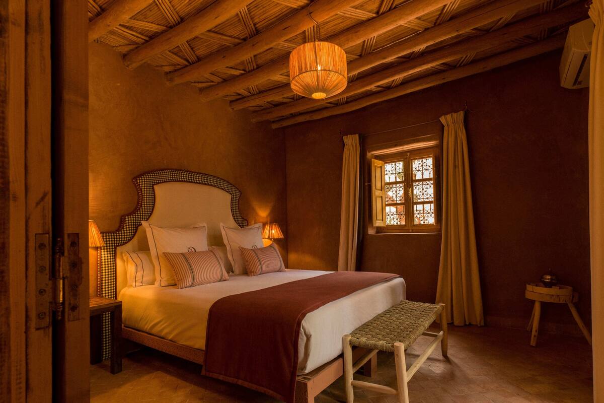 A bedroom with a wooden ceiling, a bed with a headboard, a bench, a window with curtains, and a hanging light fixture.