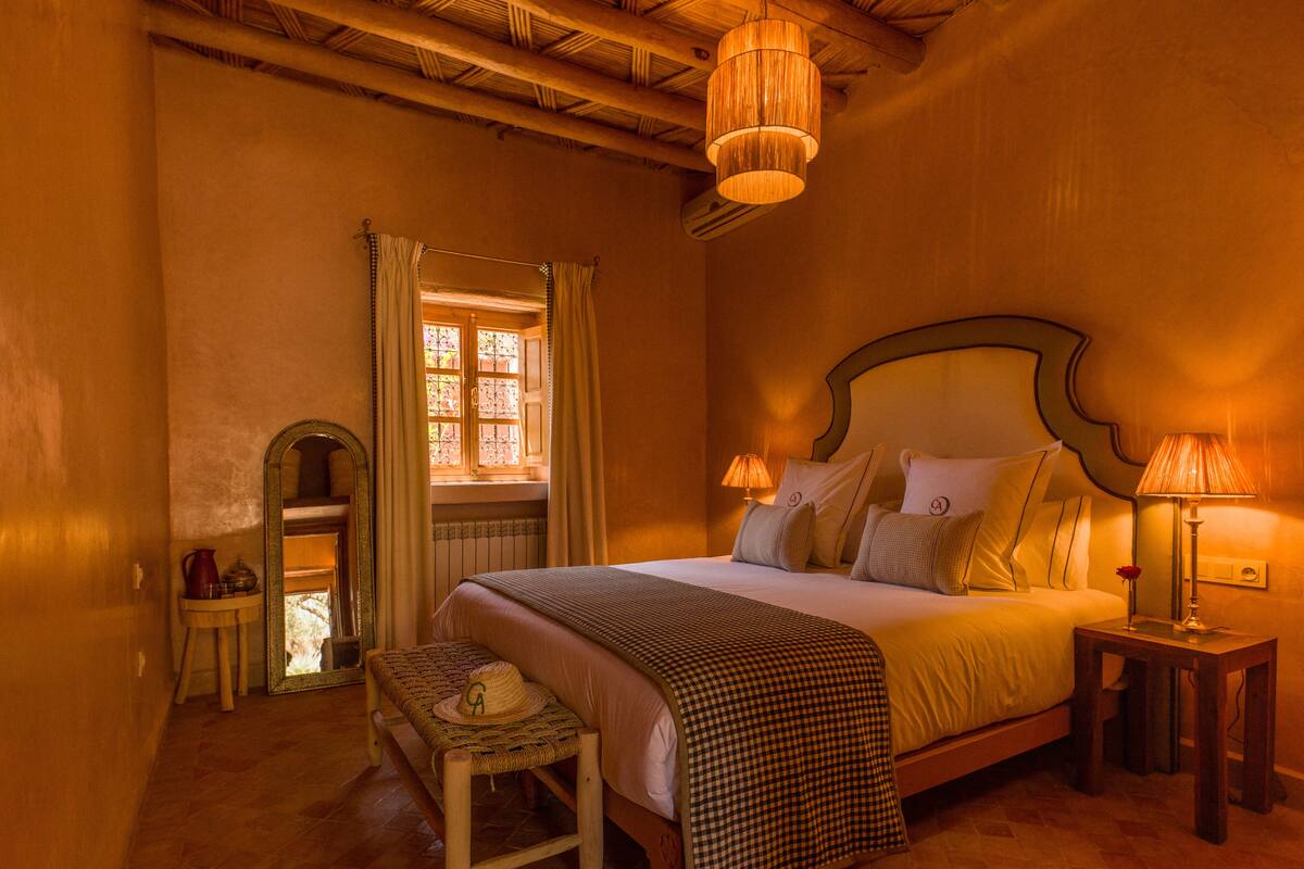 A cozy bedroom with a large bed, two bedside tables with lamps, a small bench, and a window with curtains.