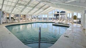 An indoor swimming pool with clear glass walls, a high ceiling, and a view of the beach.