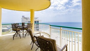 A balcony with a table and chairs, overlooking a beach with a clear view of the ocean and a building in the background.