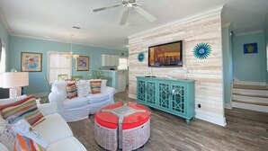 A spacious living room with a white sofa, colorful cushions, a wicker ottoman, a teal cabinet, and a ceiling fan. - Exquisite Condo Retreat, Lagoon Oasis Pool & Pristine Beaches: No Problem (Pensacola)