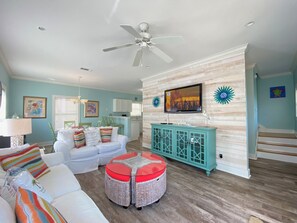 A spacious living room with a white sofa, colorful cushions, a wicker ottoman, a teal cabinet, and a ceiling fan.