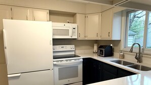 Una cocina con armarios blancos, un refrigerador blanco, un microondas, un tostador y un lavamanos con una ventana arriba.