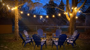 Un área de estar al aire libre con sillas Adirondack y una pequeña mesa, rodeada de árboles y luces colgantes.