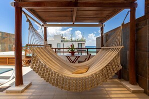 Eine Hängematte unter einer Holzpergola mit Blick auf ein Schwimmbecken und ein Gebäude gegenüber dem Wasser.