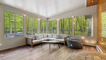 A modern living room with a sectional sofa, a round coffee table, and a bean bag chair. Large windows provide a view of a forested area.