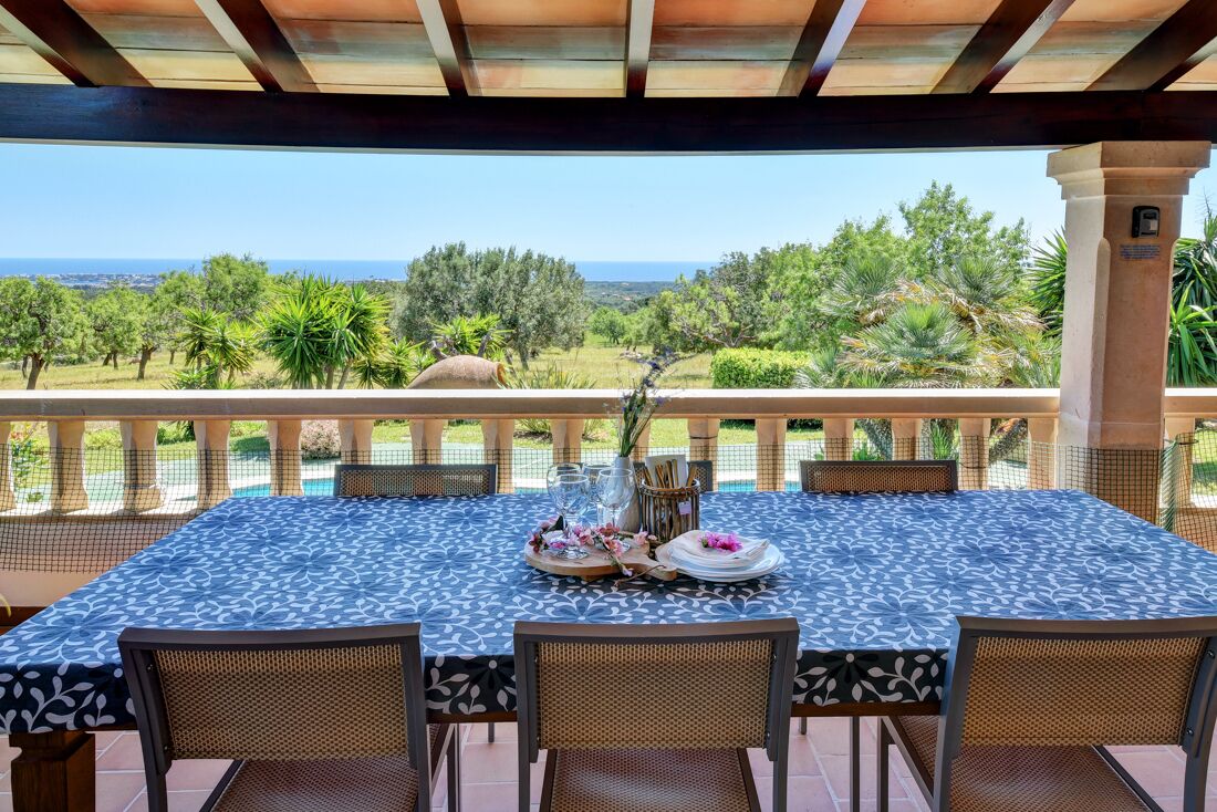 Un espace de restauration en plein air avec une table dressée pour quatre personnes, offrant une vue panoramique sur un paysage verdoyant, avec des arbres et la mer à l'horizon.