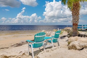 Two turquoise Adirondack chairs on a sandy beach with a palm tree and rocky outcrops.