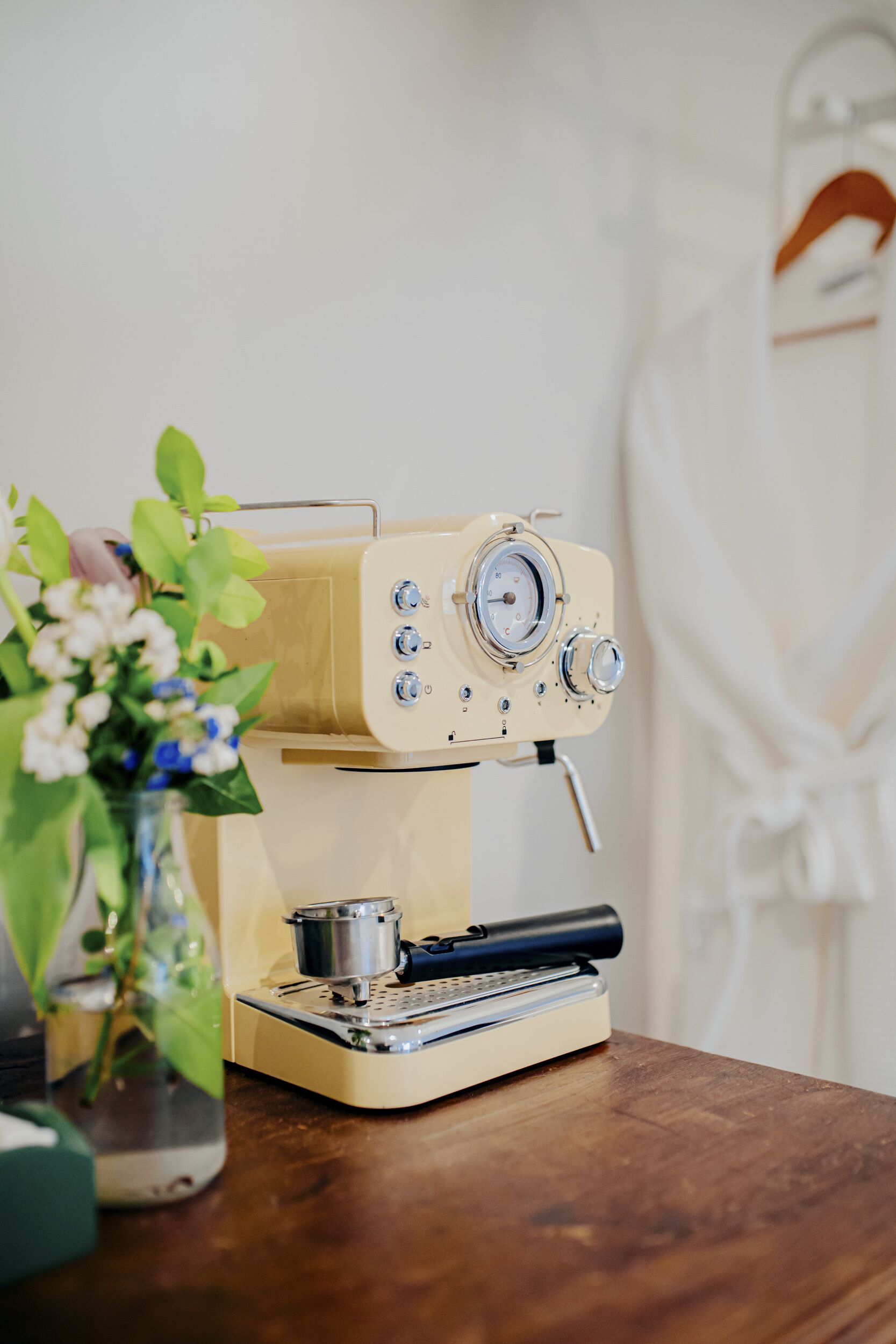 A vintage espresso machine on a wooden table with a small metal pot and a glass vase with flowers.