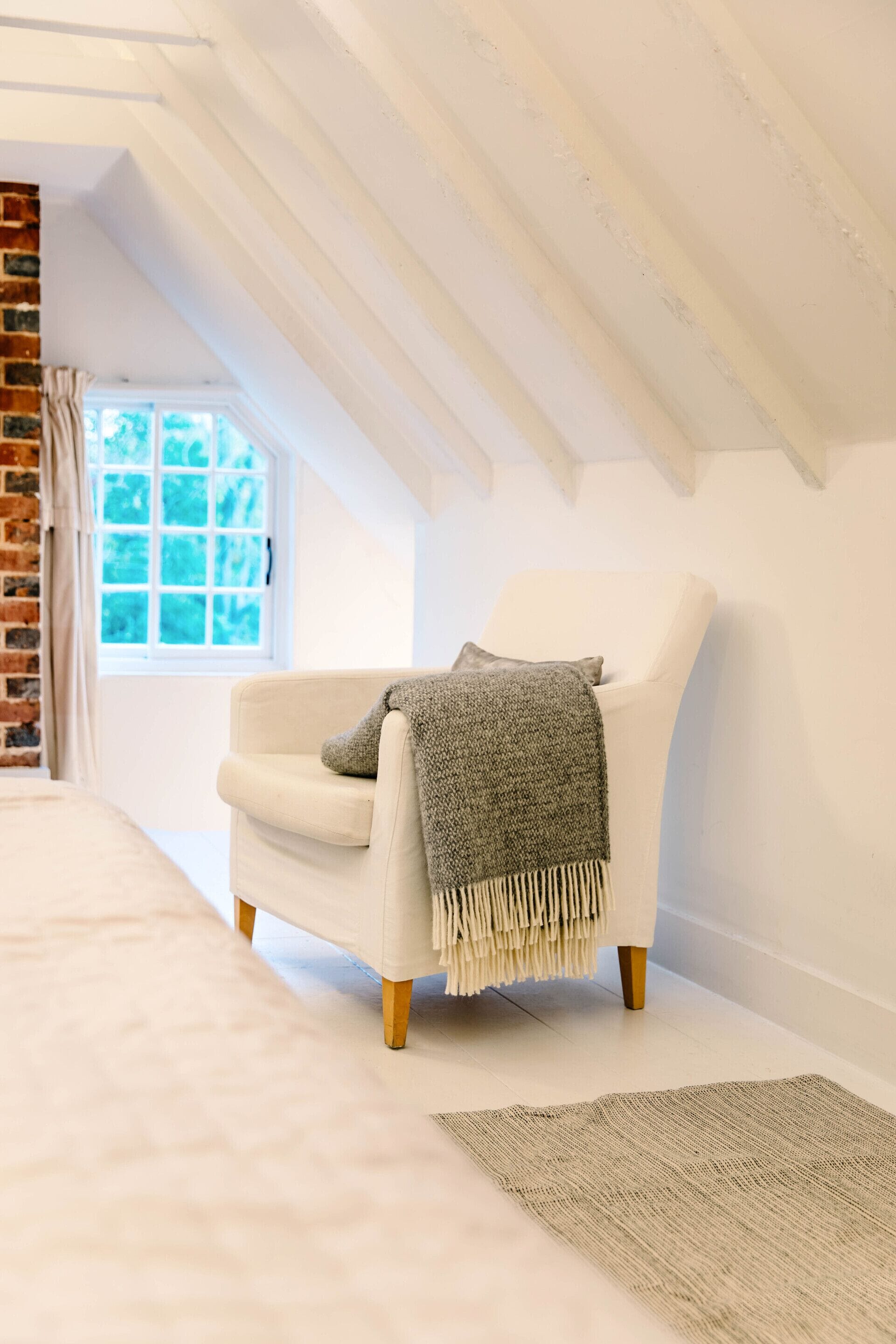 A white armchair with a grey throw blanket in a room with a sloped ceiling and a brick wall.
