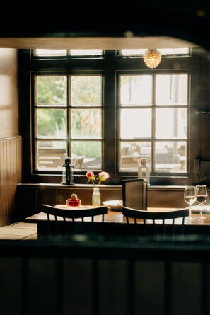 A dining area with a table set for two, a window with a view of greenery, and a decorative light fixture above the table. - The Ram Inn (Lewes)