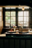 Un comedor con una mesa dispuesta para dos, una ventana con vista a follaje, y una lámpara decorativa sobre la mesa.