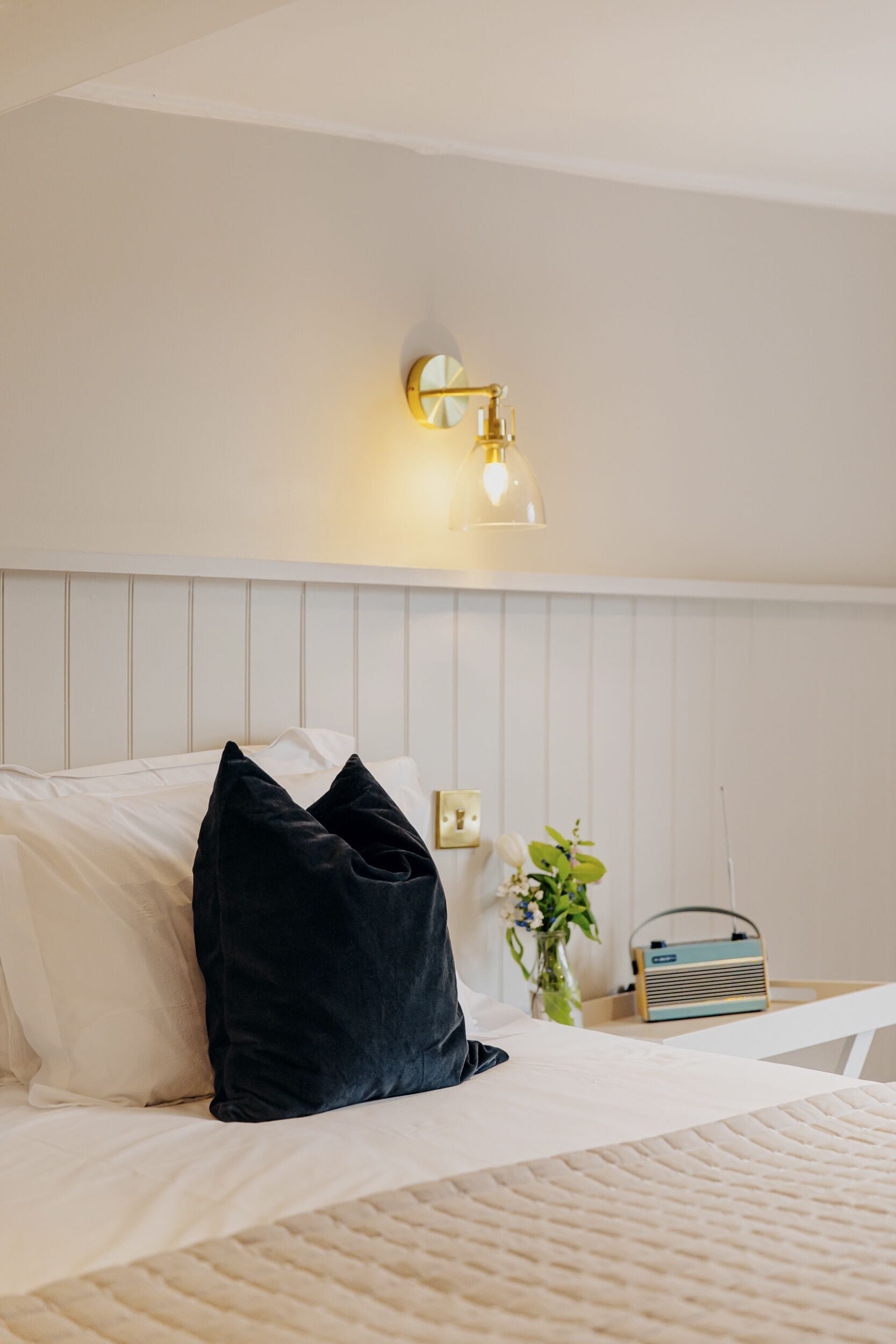 Una habitación de hotel con una cama, una mesita de noche, una lámpara, una radio y un jarrón con flores.
