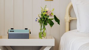 Habitación de hotel ordenada con una mesita de noche, una radio, un florero con flores y una cama con ropa de cama blanca.