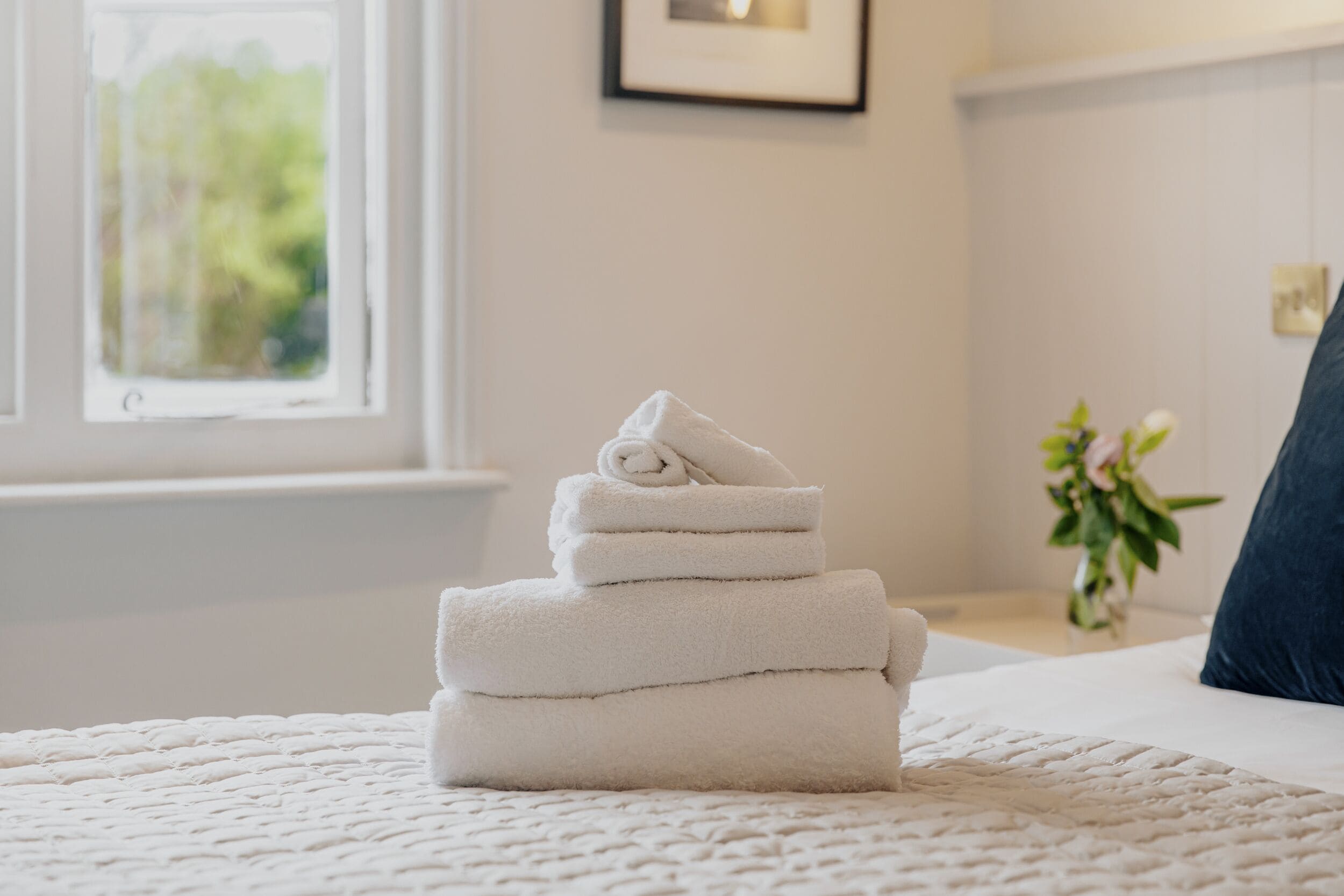 A neatly made bed with a folded white towel stack in the center, a window with a view of greenery, and a framed picture on the wall.