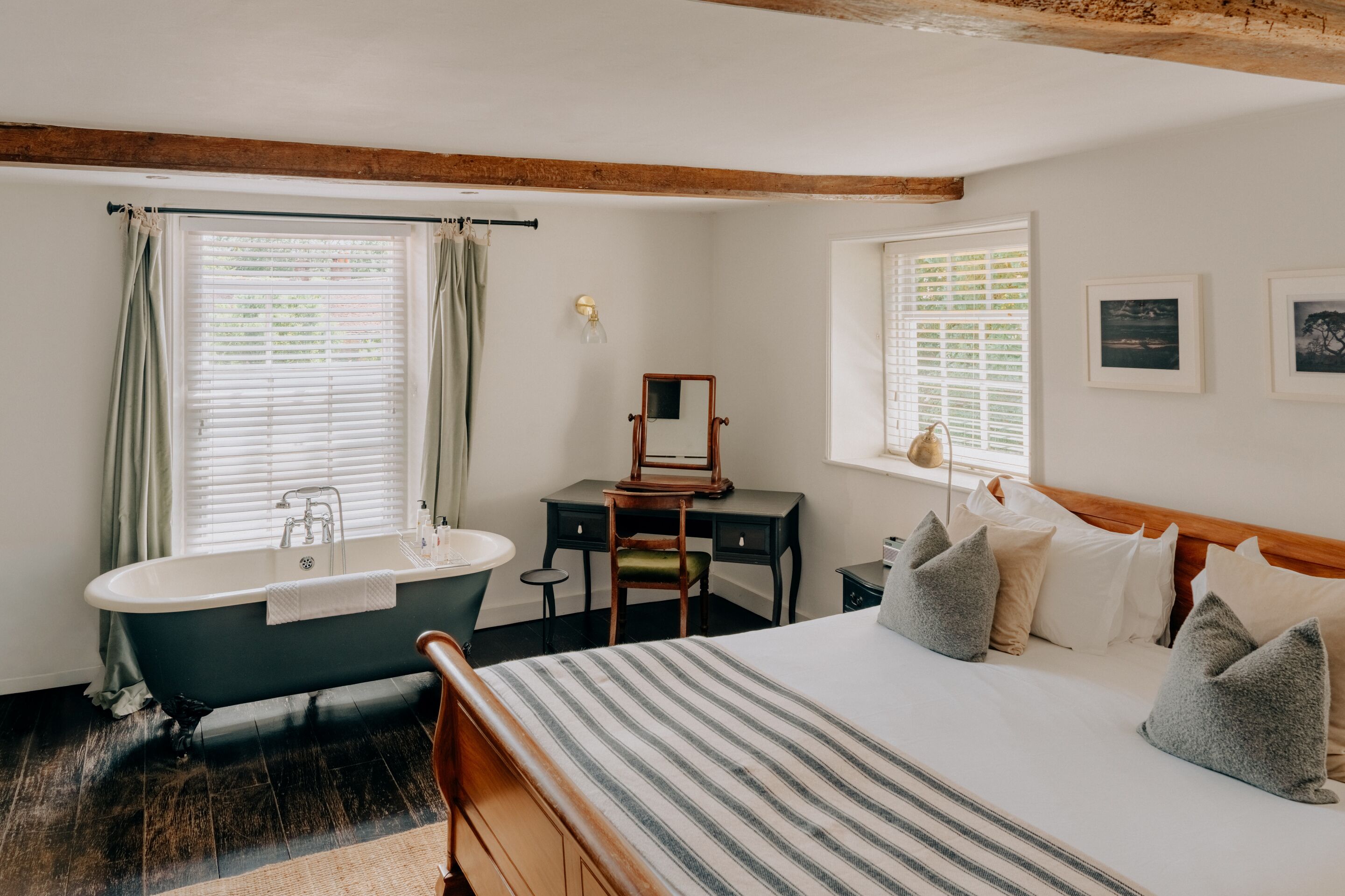 A bedroom with a large bed, a wooden headboard, a desk, a chair, a bathtub, and two windows with curtains.