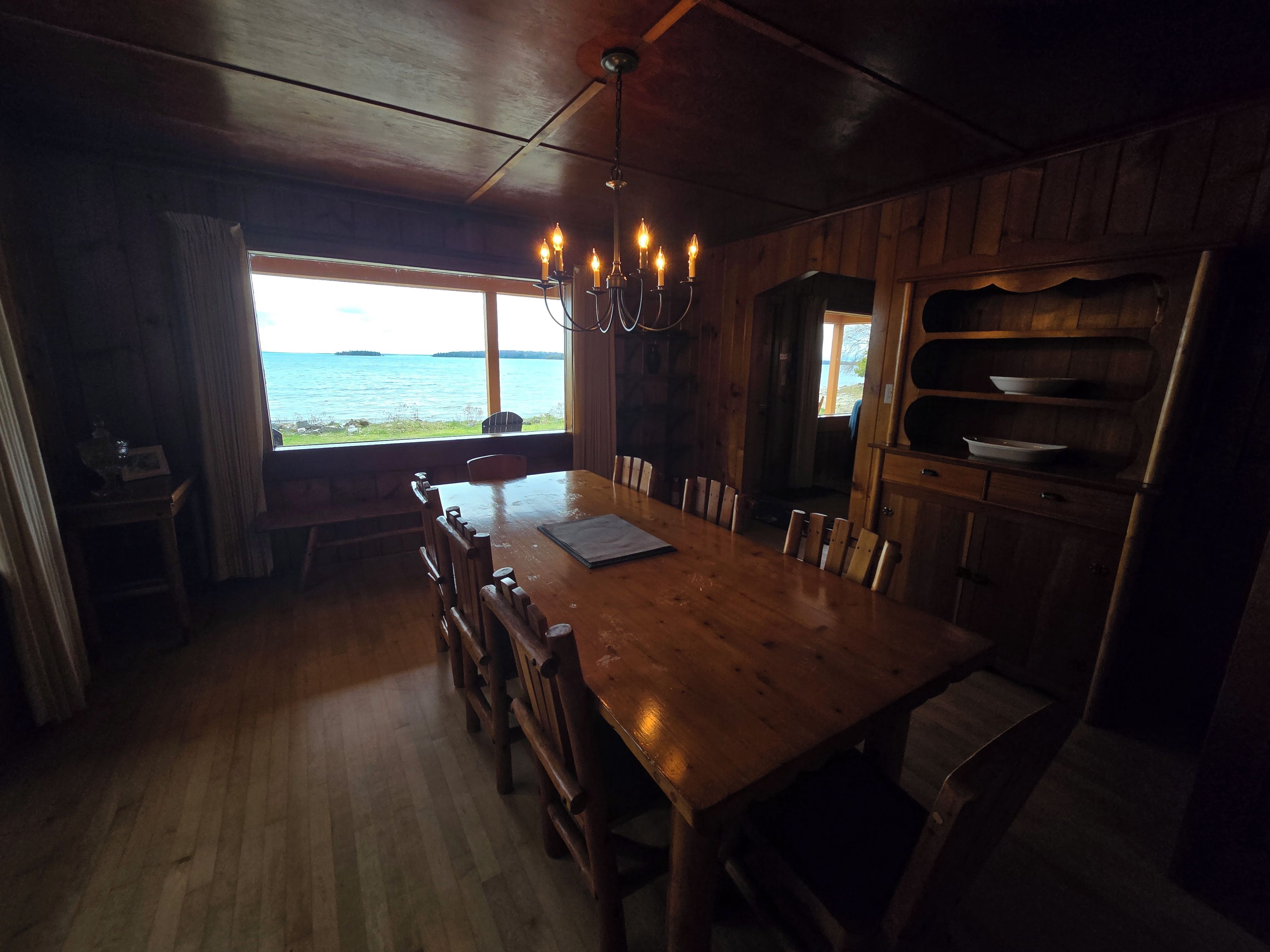 A wooden dining area with a view of the sea, a chandelier, and a wooden cabinet with dishes.