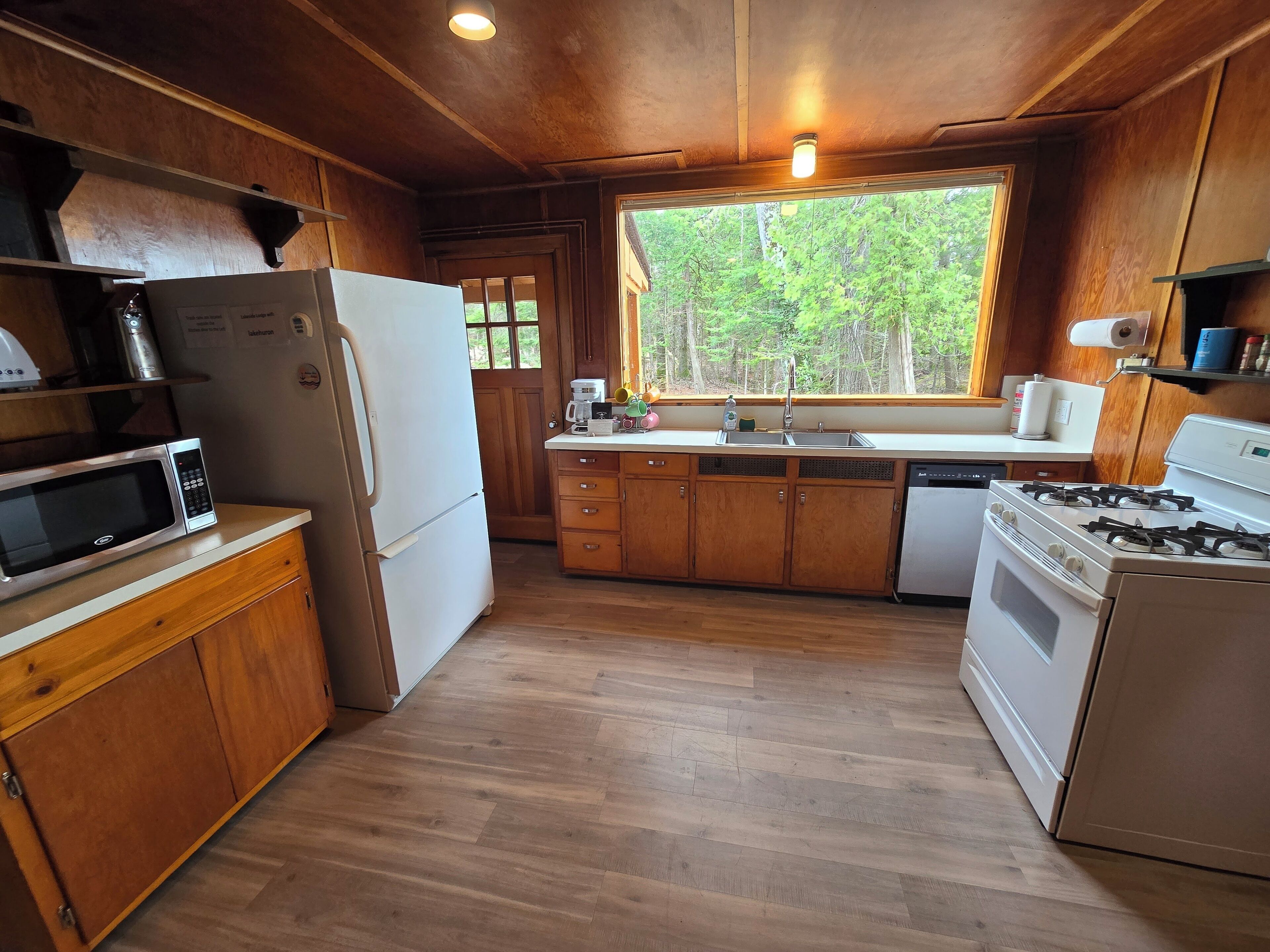 A kitchen with wooden cabinets, a microwave, a refrigerator, a stove, and a window with a view of trees.