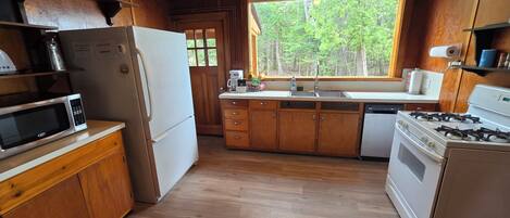 A kitchen with wooden cabinets, a microwave, a refrigerator, a stove, and a window with a view of trees.