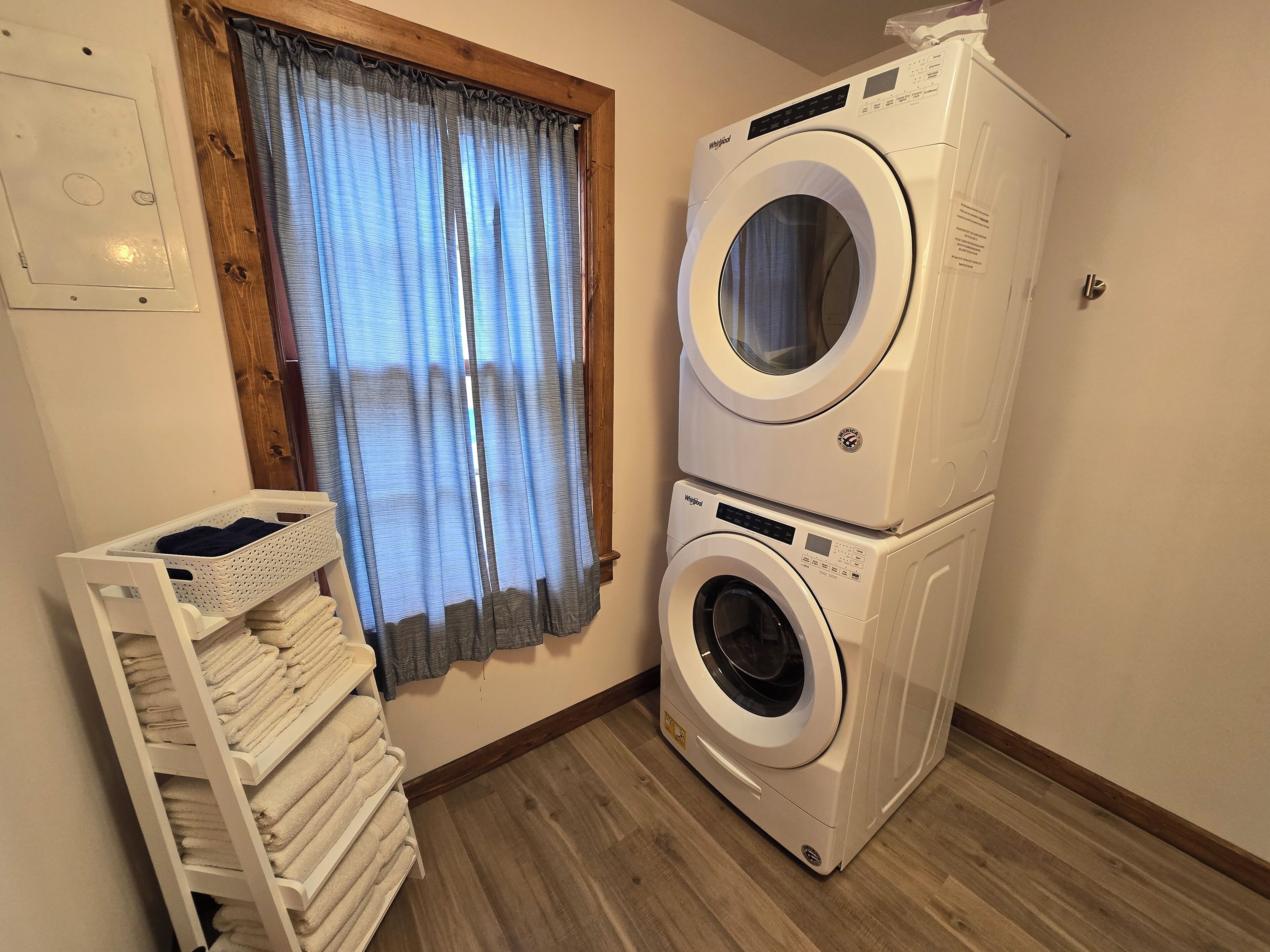 A laundry room with two stacked front-loading washing machines, a drying rack with folded towels, and a window with curtains.