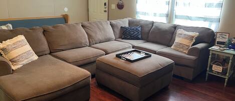 A living room with a sectional sofa, an ottoman, and a small side table with a welcome sign.