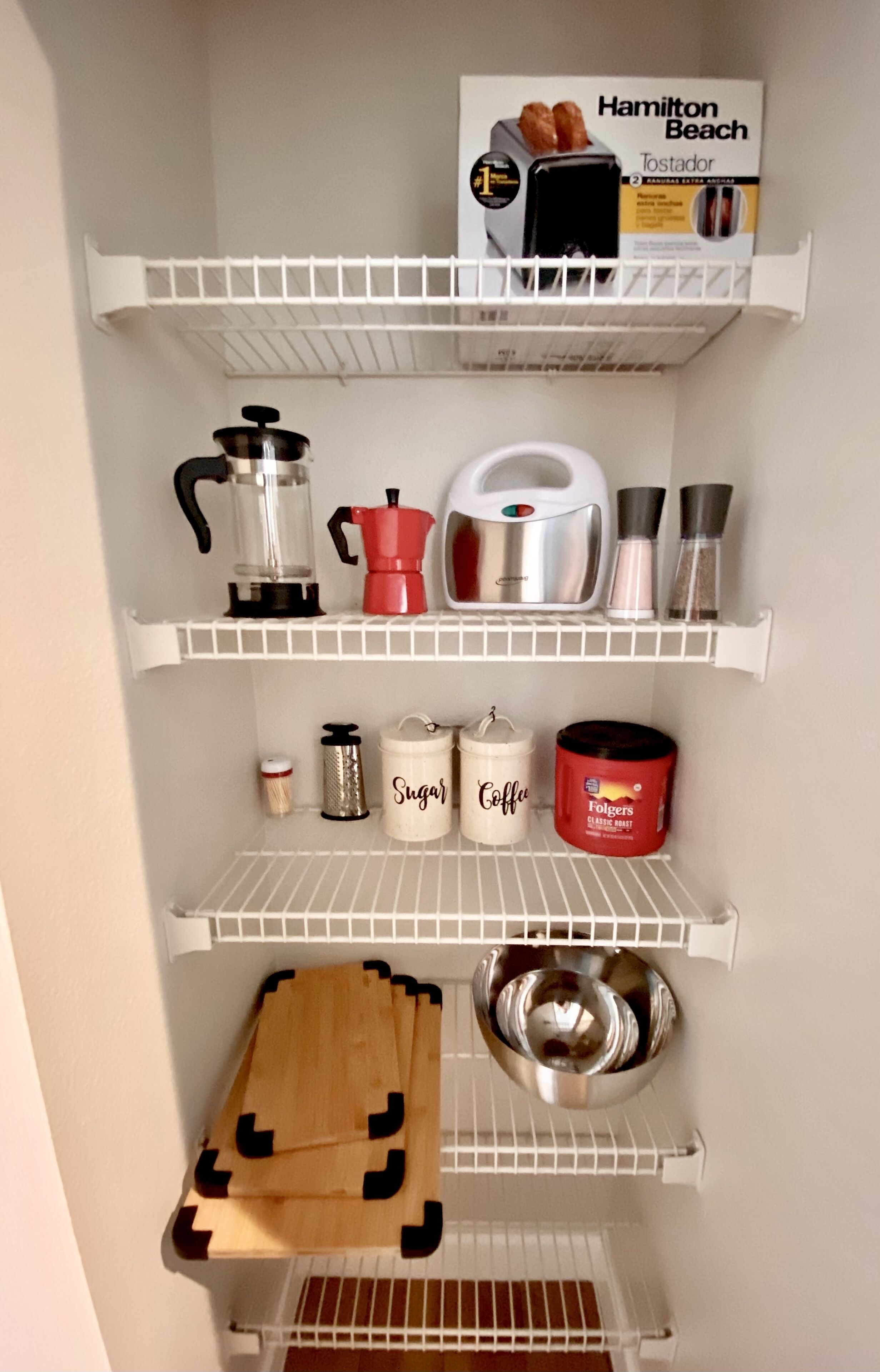Une cuisine avec des étagères blanches où sont rangés divers objets, comme un grille-pain Hamilton Beach, une machine à café et des récipients étiquetés « Sucre » et « Café ».