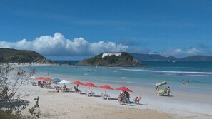 A beach with umbrellas, chairs, and a small building on a hill. - Comfortable apartment with a club and a full-service recreation area. (São Pedro da Aldeia)
