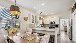 A modern kitchen with white cabinetry, a central island, and a dining area with a wooden table and chairs. - Seas The Moment- Pool- Bikes- Golf Cart- Dog Frien (Santa Rosa Beach)
