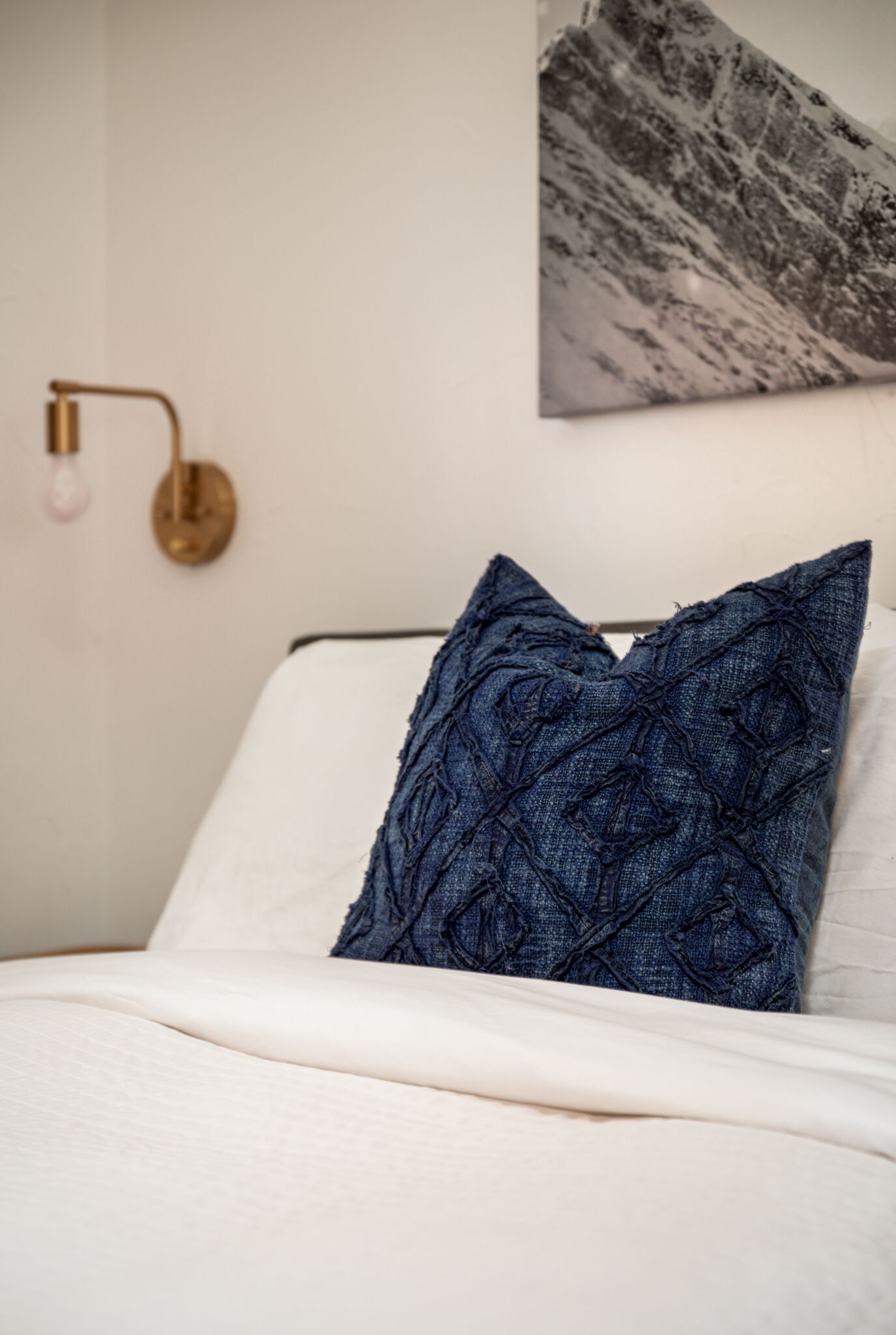 Una cama con colchón blanco y almohada azul con textura, una lámpara de pared y un cuadro en la pared.