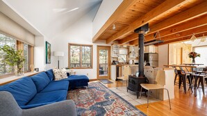 A spacious living area with a blue sofa, a gray armchair, and a patterned rug. There is a kitchen with a stove and a dining area with chairs and a table. - Holiday Hills Escape | Fire Pit | AC | Mt. Holiday (Traverse City)