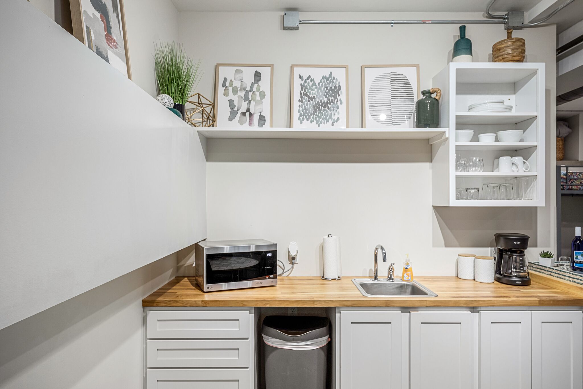 A compact kitchen with a built-in microwave, a sink, and a coffee maker. There are shelves above the counter holding various kitchen items and decorative pieces.