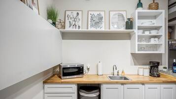 A compact kitchen with a built-in microwave, a sink, and a coffee maker. There are shelves above the counter holding various kitchen items and decorative pieces.
