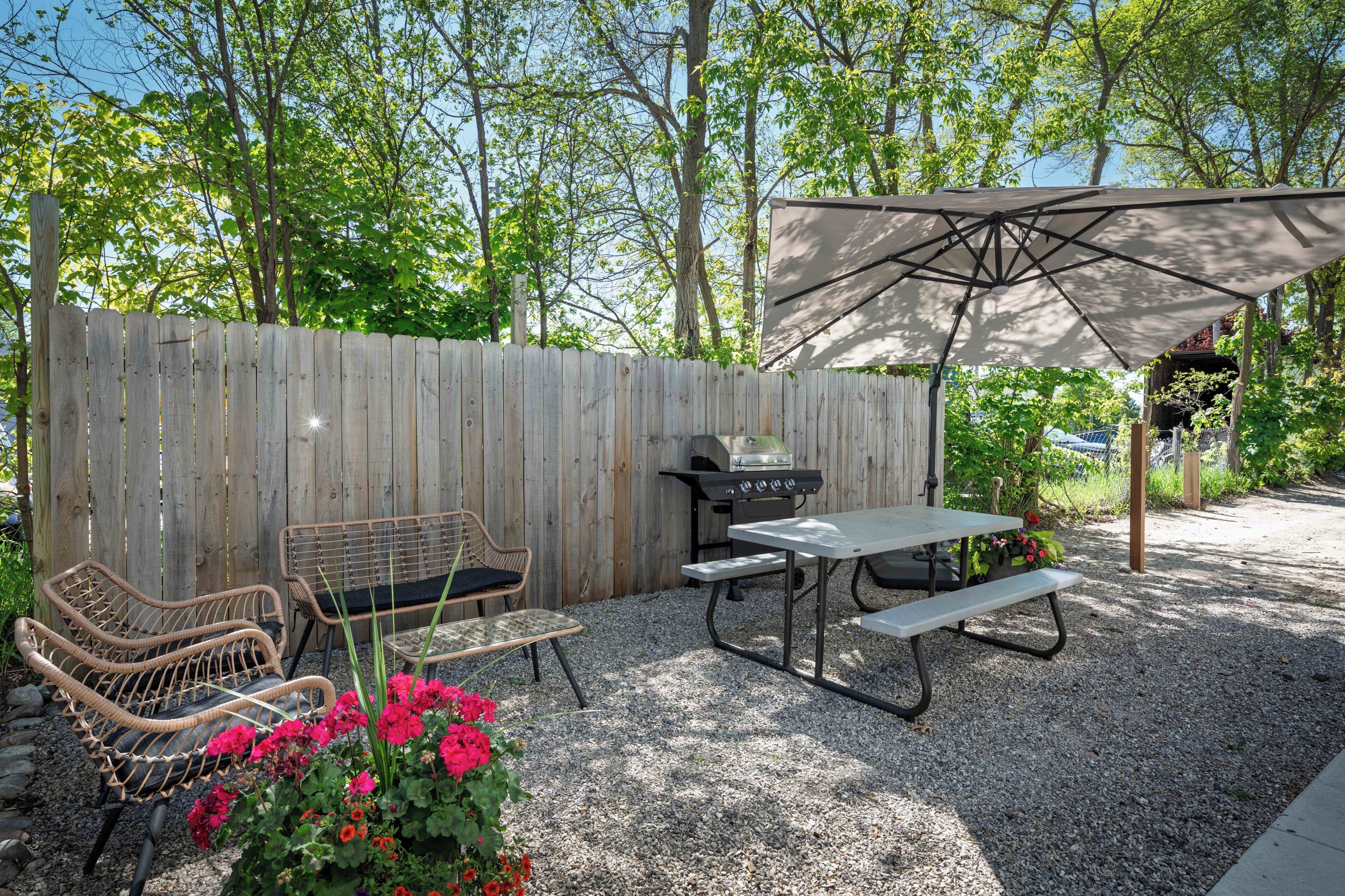 Ein Garten mit Holzzaun, Grill, Korbstühlen, Picknicktisch und Sonnenschirm.