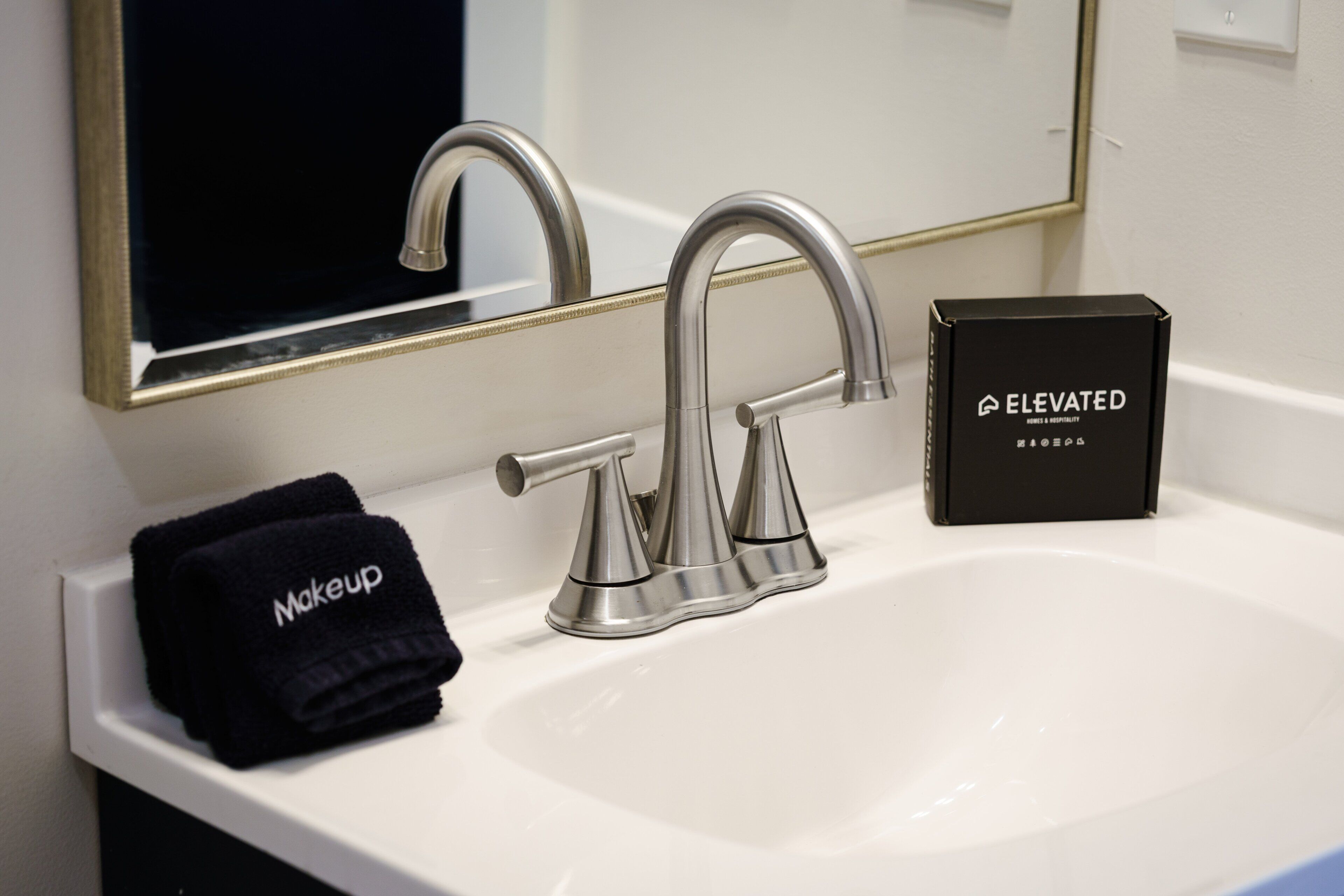 Un lavabo da bagno con un rubinetto moderno, una scatola nera con la scritta 'Elevated' e un asciugamano nero con la scritta 'Makeup'.
