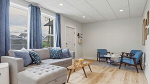 A living room with a grey sofa, blue curtains, a wooden coffee table, and a dining area with blue chairs. - Juniper @ Timberline Flats​​​​​​​ (Traverse City)