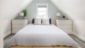 A bedroom with a sloped ceiling, a large window, a bed with striped bedding, two bedside tables with lamps and plants, and a white dresser. - Modern Mid-Century | Downtown | AC | Walkable | (Traverse City)